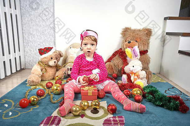 可爱的快乐小女孩基德穿着红色连衣裙，头上戴着一条鲜花做的头带，坐在车上玩圣诞玩具