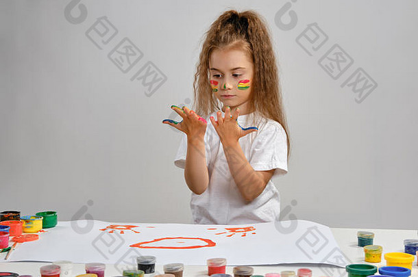 穿着白色t恤的小女孩坐在桌旁，手里拿着惠特曼和五颜六色的颜料，用手在上面画画。白色的隔离带。中等特写镜头。