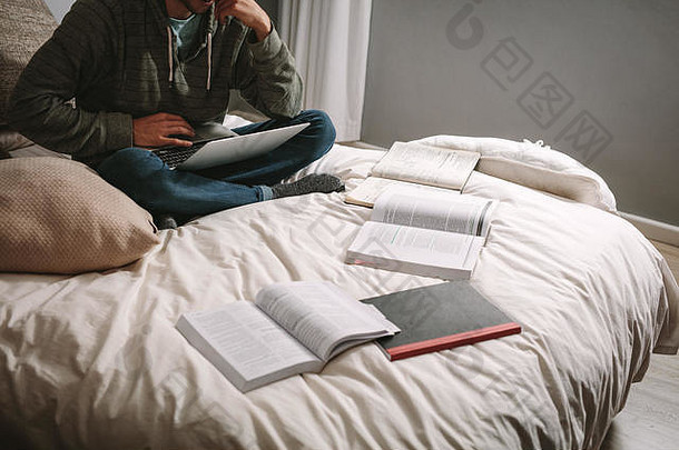 努力学习的学生坐在床上，笔记本电脑和书本放在床上。学生用笔记本电脑坐着，书摊在床上。