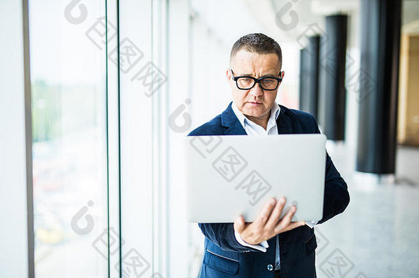 站在办公室里，一个穿着正装的开朗的成熟男人，一边调整眼镜，一边拿着笔记本电脑