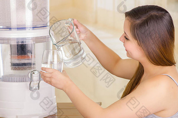 美丽的女人一手拿着一杯水，另一手拿着一罐水，厨房背景上有一个<strong>净水器</strong>过滤系统