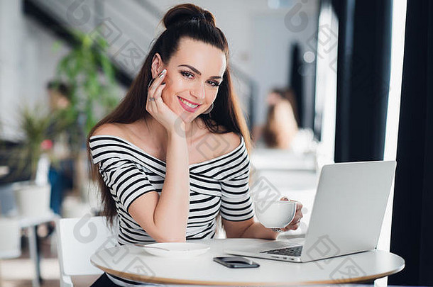 微笑的成年女子坐在咖啡馆里，拿着笔记本电脑，端着一杯咖啡，看着摄像机。