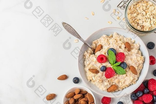 一碗燕麦片粥，配上新鲜浆果、杏仁和薄荷，在白色餐桌上享用健康饮食早餐。具有空间的俯视图