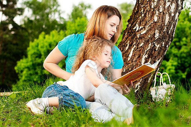 年轻的母亲坐在树下的草地上，给躺在母亲膝上微笑着的小女儿读书