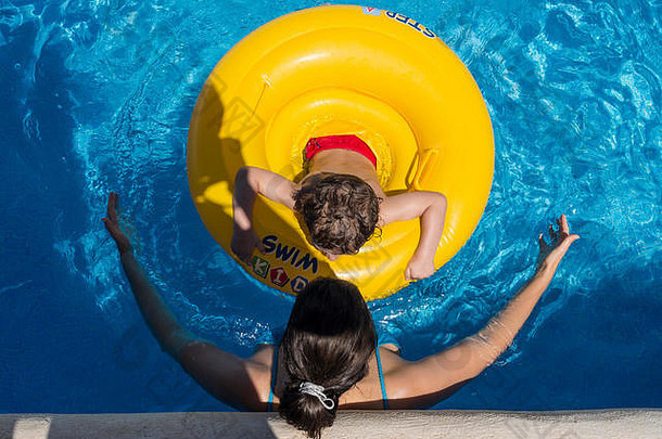 游泳池里，一个孩子带着一个黄色的漂浮物，旁边是一个女人，水中倒影。顶视图。