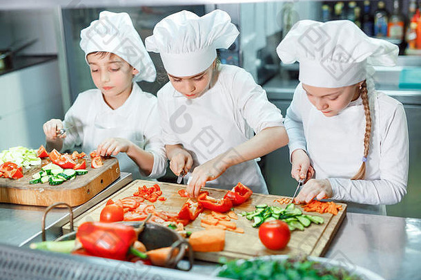 戴厨师帽的孩子们在厨房里磨蔬菜。