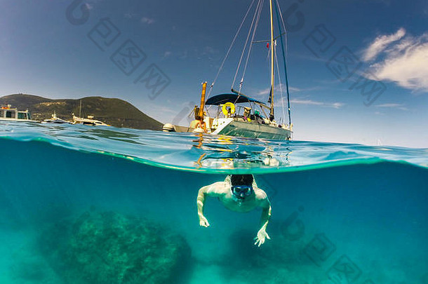 一名男子在清澈的蓝色海洋中游泳，背景是一艘游艇，一半在水面上，一半在海里，这张<strong>照片</strong>是用圆顶相机拍摄的