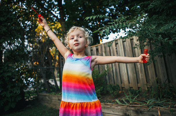 夏日日落时，可爱有趣的金发白人小女孩在自家后院吹肥皂泡的真实写照。真正的快乐童年