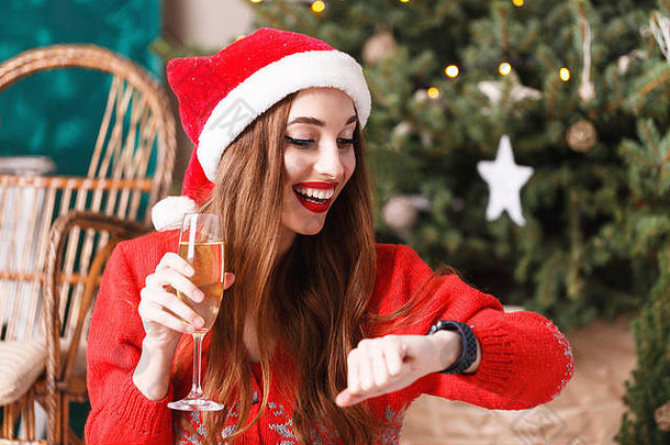 穿着圣诞帽和红色毛衣的迷人圣诞妇女坐在舒适的新年云杉旁的地板上，拿着香槟等待新年，她看着手表、新年、圣诞节、假日、纪念品、礼物、购物、折扣、商店、雪人圣诞老人、化妆、发型、狂欢节。