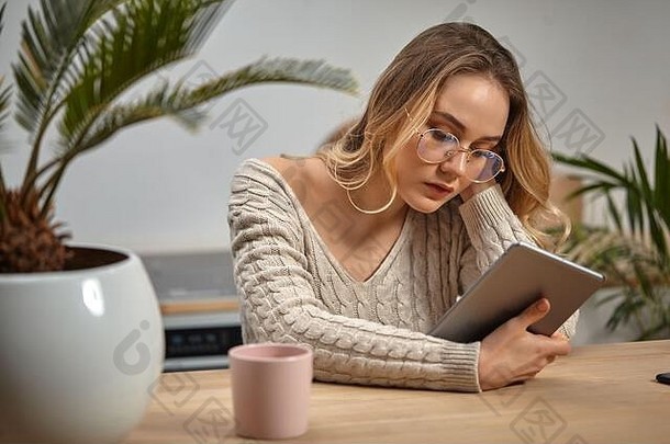 金发女郎女孩博主眼镜米色针织毛衣持有平板电脑坐着厨房木表格棕榈树粉红色的杯特写镜头