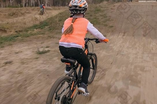 一名白人儿童在土公园的自行车道上玩耍。在赛马场骑<strong>黑橙</strong>色自行车的女孩。这孩子去做自行车运动。骑自行车的运动骑乘