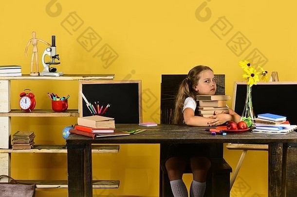 脸色暴躁的女学生手里拿着一摞书。黄色背景上的<strong>儿童</strong>和学校用品。回到学校和教育理念。女孩坐在木制书桌旁，桌上放着五颜六色的文具、鲜花和水果