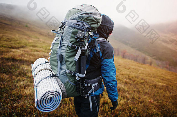 背部游客背包垫和三脚架的特写镜头，穿着带风帽的蓝色旅游夹克，站在山上