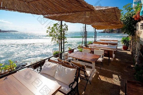 海边一家餐馆的空阳台。晨光中海湾的美丽景色。木制桌子和座椅上方的装饰伞。晴朗的天气