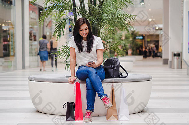 一位女士坐在商场里，看着她的手机，微笑着，地上放着一些购物袋。购物理念。