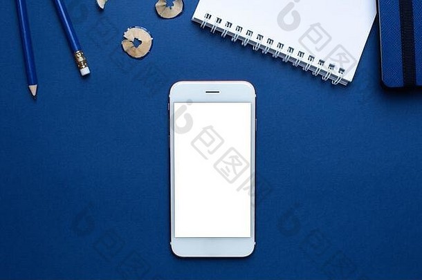 手机和白色、蓝色的办公笔记本，铅笔放在经典的蓝色背景顶视图上。时尚潘通颜色2020