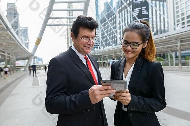 快乐成熟的商人和年轻的亚洲女商人在城市里使用数字平板电脑