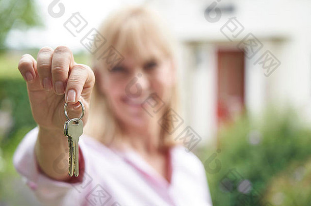 一幅成熟女子手持钥匙站在乡村<strong>梦想家园</strong>前的花园中的画像