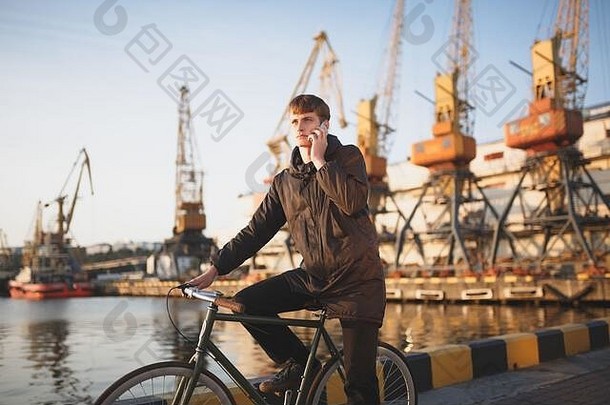 一个棕色头发的年轻人站在自行车旁，一边说着手机，一边若有所思地向一旁看。穿羽绒服骑自行车的<strong>酷男孩</strong>
