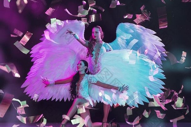 年轻的女舞者，带着白色天使的翅膀，在紫色蓝色霓虹灯下，黑色背景下，五彩纸屑飞舞。优雅的女子舞蹈。狂欢节的概念，美，运动，克服，绽放。