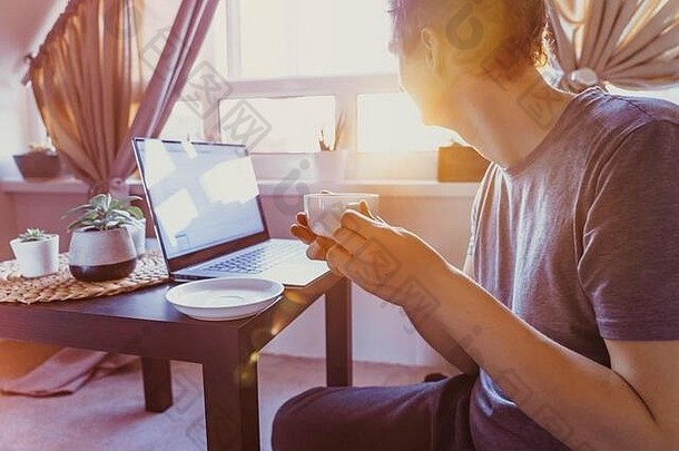 坐在咖啡桌旁用笔记本电脑工作的年轻人，端着一杯热饮茶，欣赏日落美景。工作<strong>场</strong>所靠近窗户。在家工作。<strong>返</strong>回