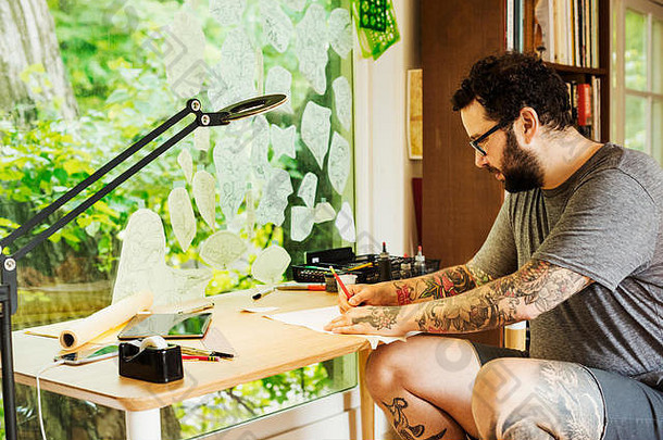 一个留着胡子、手臂和腿上都有纹身的男人坐在桌子旁画画。