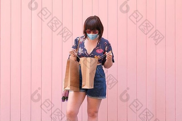 年轻的高加索人女人穿脸面具太阳镜内部购物袋粉红色的背景冠状病毒购物概念