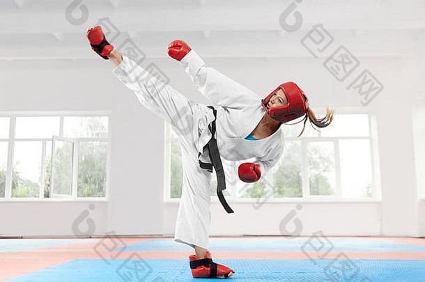 身穿传统白色和服和黑带的年轻女子在光明课堂上练习空手道动作。穿着红色防护手套和头盔的运动型女孩，穿着展示武术踢脚的鞋子。