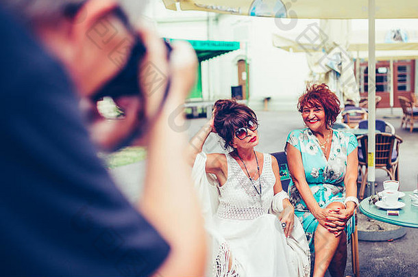摄影师与坐在室外咖啡馆的两位快乐的老年妇女合影。