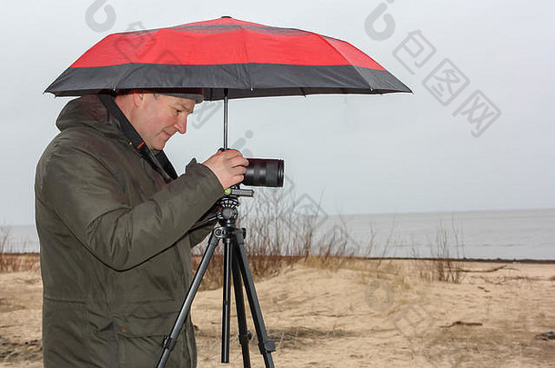 一个年轻人拿着一把红伞，在下雨天拍海滩照片。