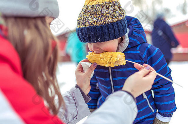 一个小男孩在冬季集市上吃煮熟的玉米，周末在大自然里吃，蓝色保暖连体衣，保暖帽子，妈妈喂孩子，早餐和午餐都吃零食