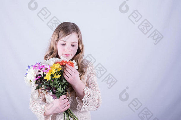 一个留着又长又脏的金发的12岁女孩看着一束五颜六色的雏菊，手里拿着一朵橙色的雏菊，背景是白色的
