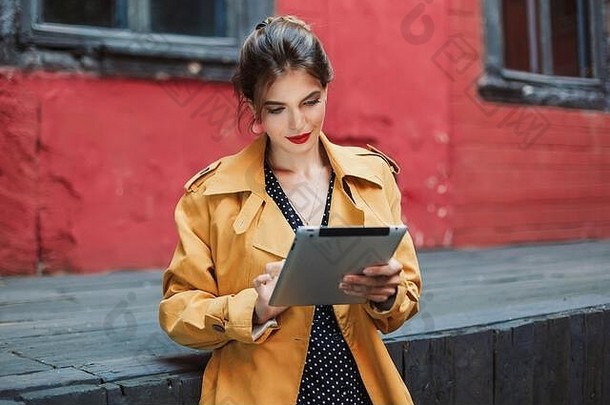 穿着橙色风衣和黑色圆点衣服的年轻迷人的女人在老院子里度过时光时，若有所思地使用平板电脑