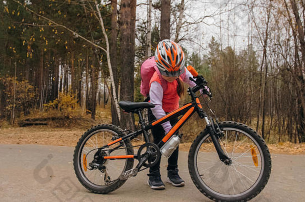 一个白人孩子在秋天公园里骑自行车散步。小女孩在森林里骑着<strong>黑橙</strong>色的自行车。这孩子去做自行车运动。带背包和头盔的自行车运动骑乘。山地车硬尾。