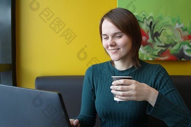 女孩快乐的表达式脸微笑坐着移动PC表格咖啡馆棕色（的）沙发黄色的墙电脑监控持有纸杯咖啡手