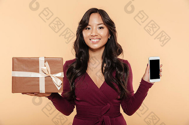 身着栗色连衣裙的亚洲微笑女子<strong>展示礼品</strong>盒，智能手机作为礼物，在桃色背景下被孤立