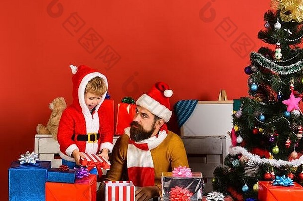 一个留着胡子、脸上带着兴趣的男人和他的儿子玩。圣诞老人和小精灵在冷杉树附近的礼品盒中。圣诞家庭在红色背景上打开礼物。寒假与庆典理念