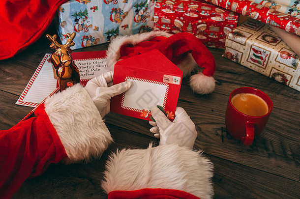 戴着白手套的圣诞老人在他的木桌旁拿着一个信封，里面有礼物、包装纸、圣诞帽、装咖啡的红杯子和通讯员