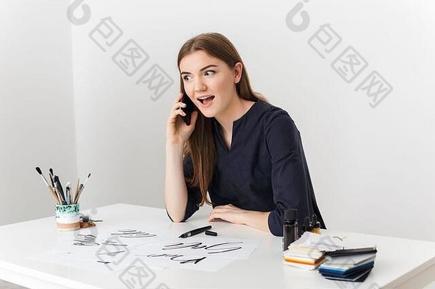 一张年轻漂亮的女士坐在白色书桌旁，一边用手机聊天，一边愉快地向一边看的照片