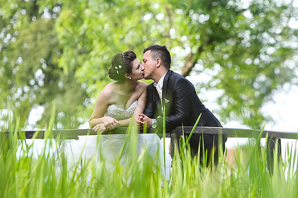 新娘和新郎站在一座木桥上，倚在篱笆上接吻。