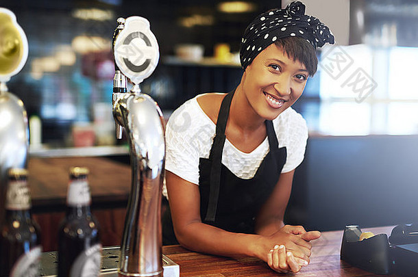 迷人的年轻非洲裔美国小企业主，戴着头巾和围裙，靠在酒吧的柜台上