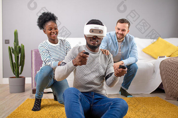 快乐微笑的多文化朋友尝试3D虚拟现实护目镜。一个非洲男人在家玩虚拟游戏，而他的朋友们则在家玩游戏