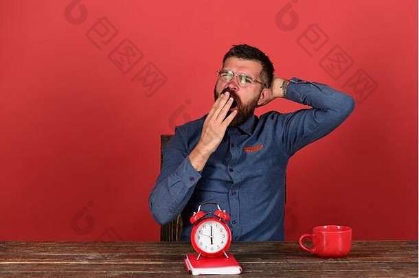 最后期限和工作理念。面容疲惫的男子坐在木桌旁。留着胡须、戴着眼镜、打哈欠的工人，浅红色<strong>背景</strong>。茶杯、复古时钟和红皮书放在复古桌上