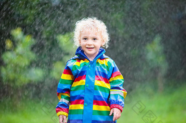 金发卷发的小男孩穿着彩虹色的防水夹克在雨中玩耍。孩子们在户外享受秋雨的乐趣。