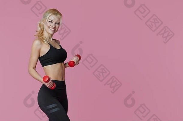 健身女孩，完美修长的身体训练肌肉，粉色背景上有哑铃。穿着运动服的运动型女人