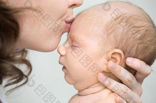 有趣的新生婴儿在妈妈怀里打盹。年轻的母亲亲吻着睡着的可爱的新生儿的额头。健康小