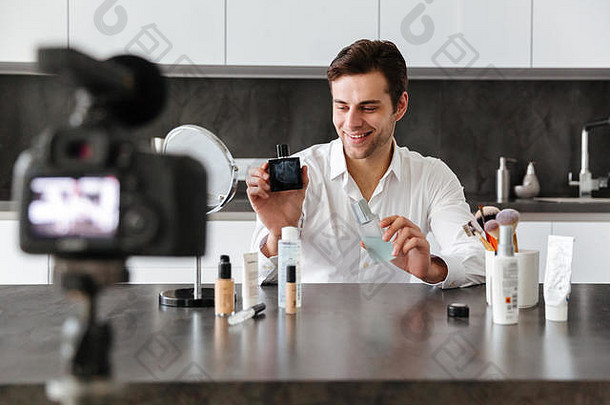 一个快乐的年轻人坐在厨房的桌子旁，一边化妆，一边在他的视频博客上拍摄关于新化妆品的插曲