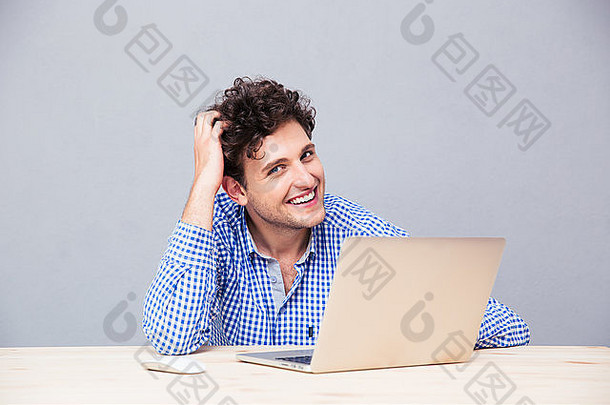 快乐的男人坐在桌子旁，把笔记本电脑放在灰色背景上，看着摄像机