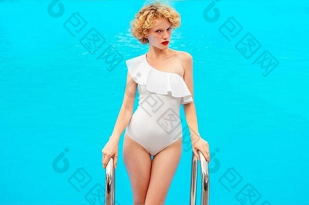 美丽的红发卷发女人，穿着白色时尚的游泳衣，站在夏日室外的游泳池楼梯上。夏天，快乐的概念