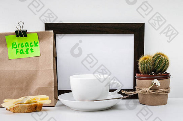 模拟的深色相框、仙人掌、一个印有早餐字样的纸袋、一个奶酪三明治和一个白色杯子，在浅色背景上放着一碟咖啡